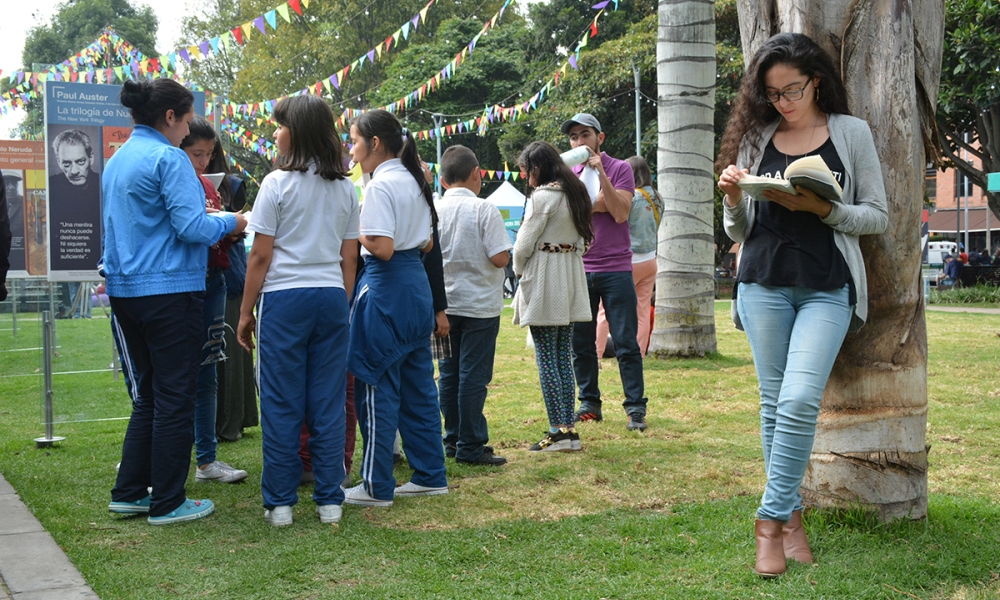 Mujer leyendo libros mientras se recuesta en un árbol, junto a ella hay unos niños con uniforme realizando una actividad en un parque