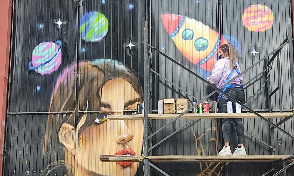 una mujer sobre un andamio pinta la fachada de un Centro de Formación. El grafiti consiste en un rostro de mujer y una nave espacial.