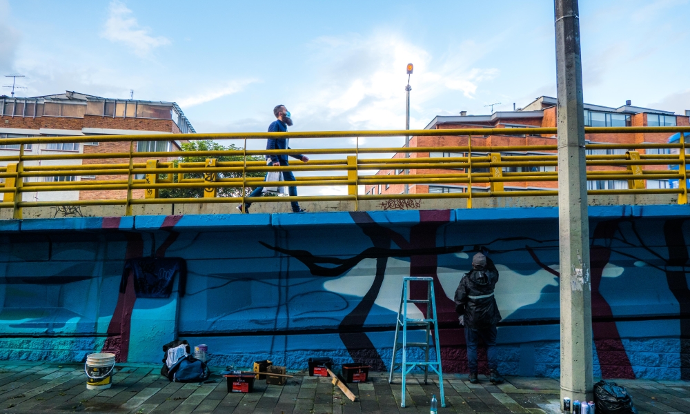 Artista urbano realiza su intervención en un puente de Bogotá mientras un hombre lo cruza