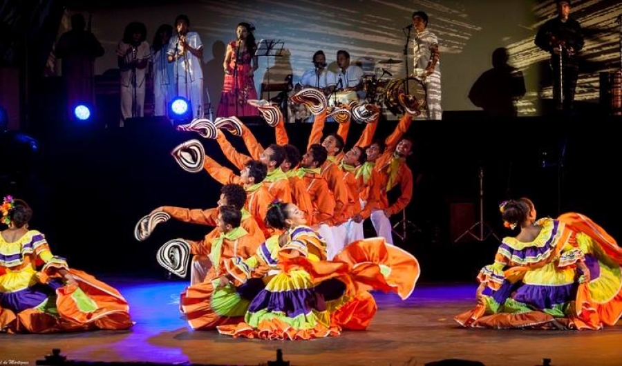 Grupos debailarines vestidos de colores en escenario con grupo musical al fondo.