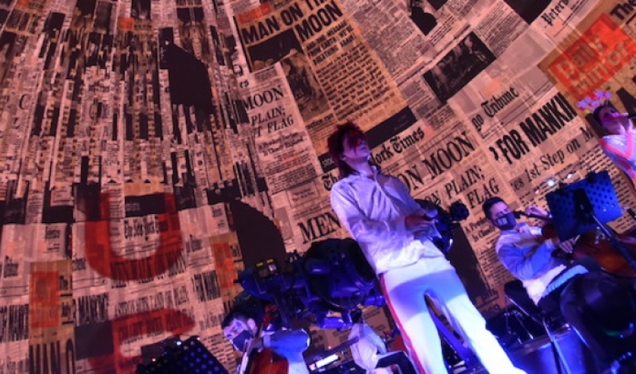 Hombre vestido como David Bowie en la tarima del domo del Planetario con imágenes de periódicos en el fondo. 