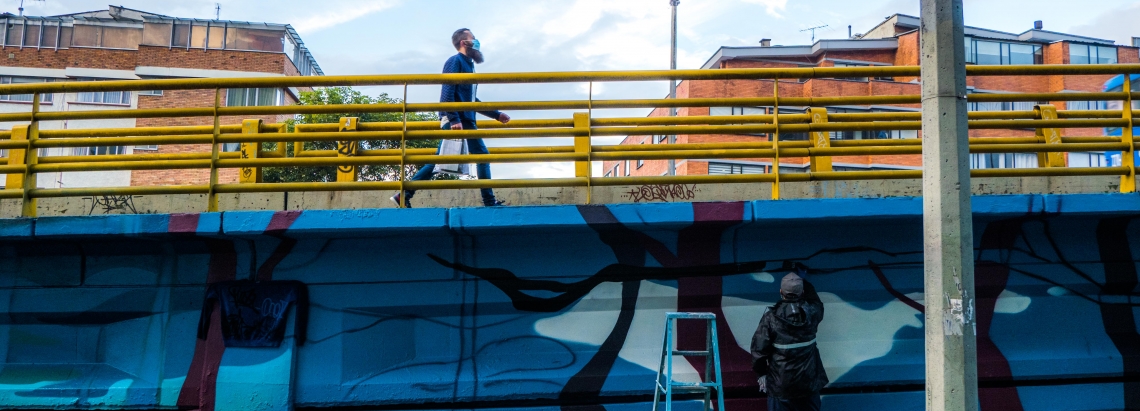 Artista urbano realiza su intervención en un puente de Bogotá mientras un hombre lo cruza