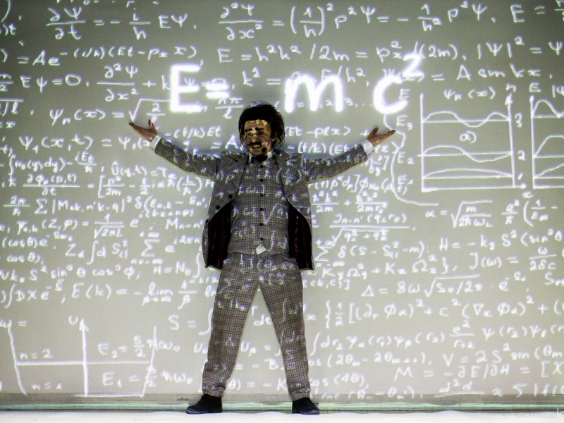 Imagen de actor representando a Albert Einstein al frente de un tablero lleno de formulas matemáticas