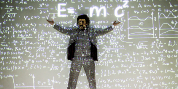 Imagen de actor representando a Albert Einstein al frente de un tablero lleno de formulas matemáticas