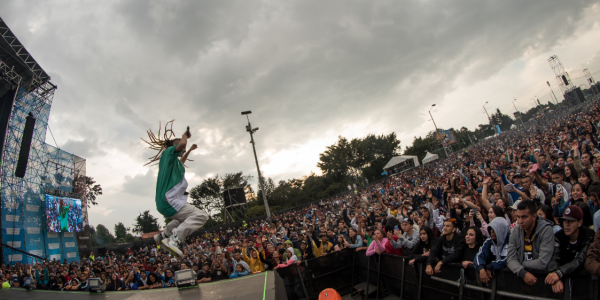 Hombre de pelo largo saltando en la tarima de un festival de música ante un público masivo.
