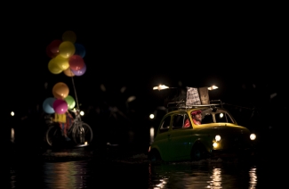 Auto amarillo con equipaje y conductor y hombre en bicicleta con globos de colores sobre el lago.