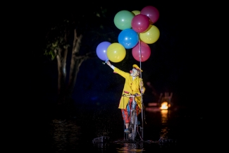 Payaso de rojo y amarillo en bicicleta con globos sobre el lago.